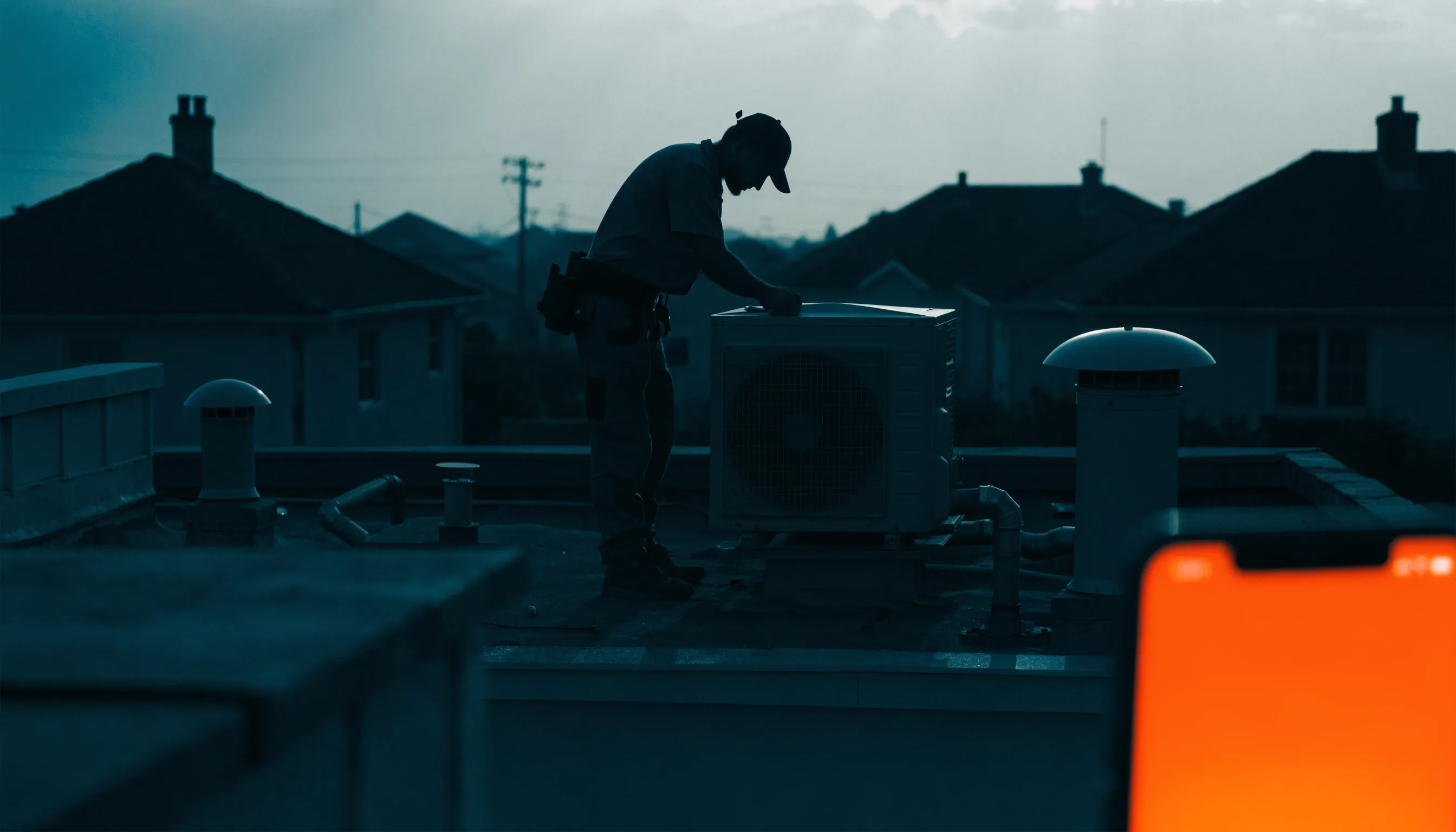 An air-con technician silhouetted on a rooftop at dusk with a phone glowing orange