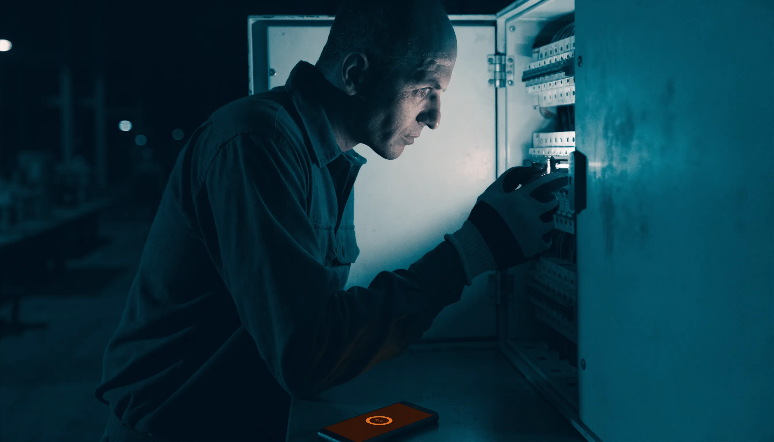 An electrician working at a switchboard at night with a phone glowing orange nearby
