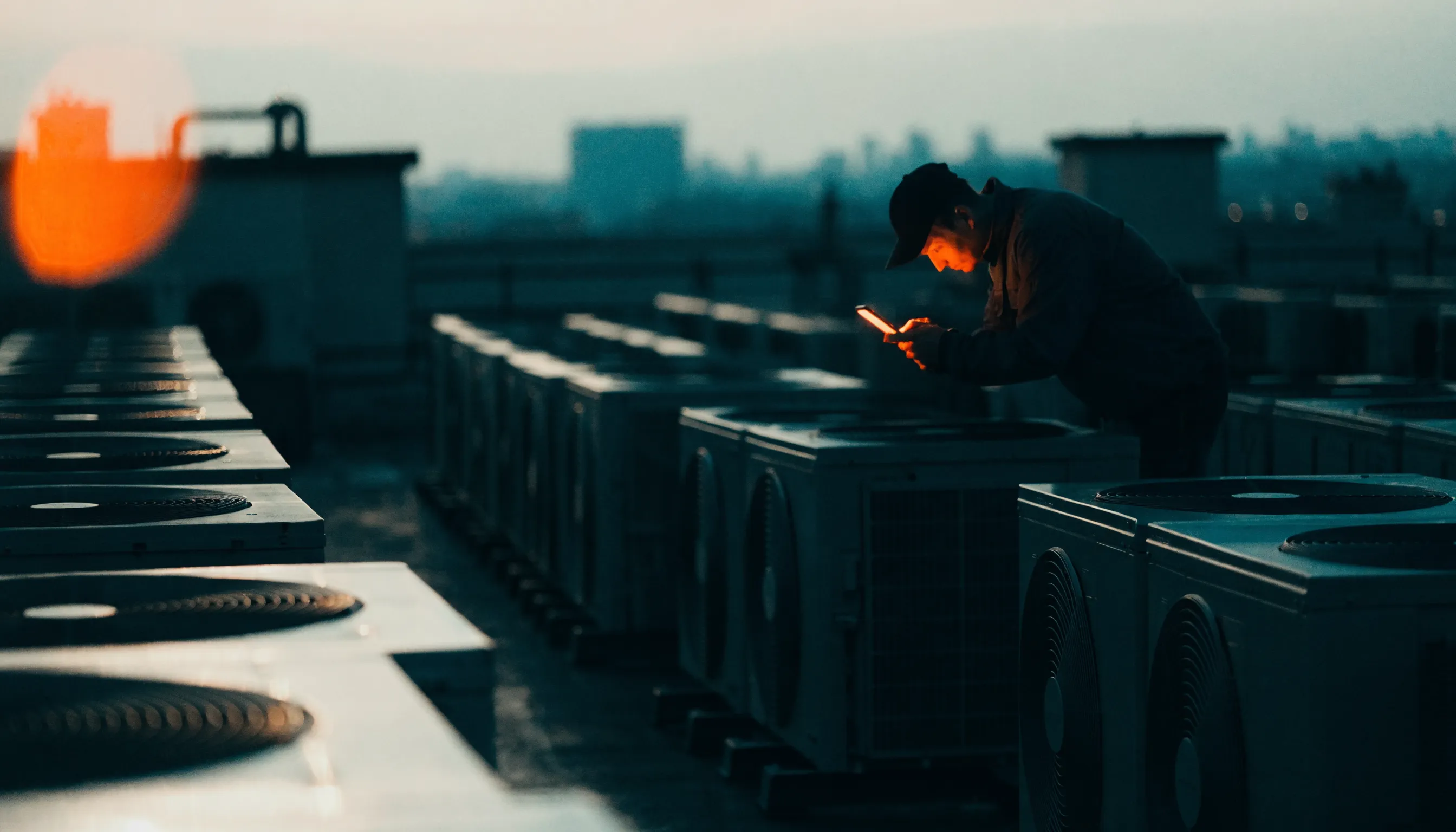 Rows of air-conditioning units on a rooftop at dusk with a technician holding a phone glowing orange