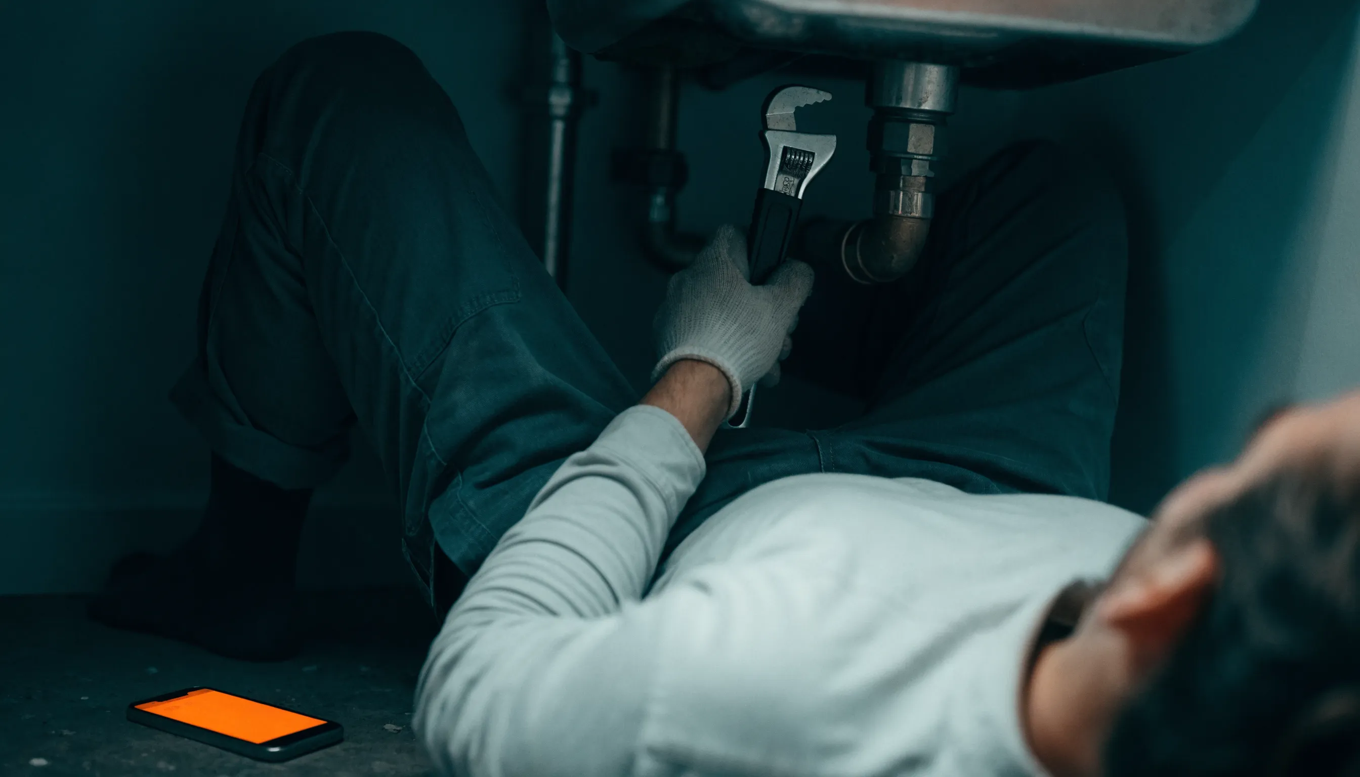 A tradesperson working under a sink with a phone glowing orange on the floor, out of reach